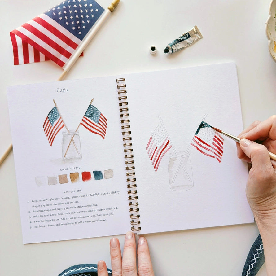 Person painting an American flag in a sketchbook with a small American flag and paint tube on a white surface.