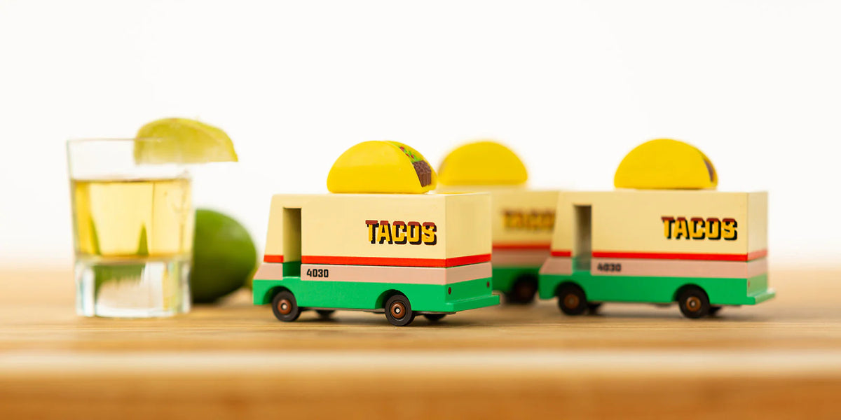 Toy taco trucks with 'TACOS' branding on a wooden surface with a glass of limeade.