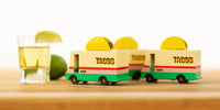 Toy taco trucks with 'TACOS' branding on a wooden surface with a glass of limeade.