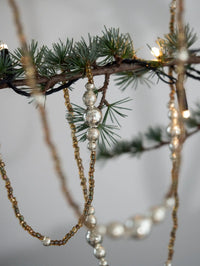Decorative beaded strands hanging from a branch with small lights against a neutral background