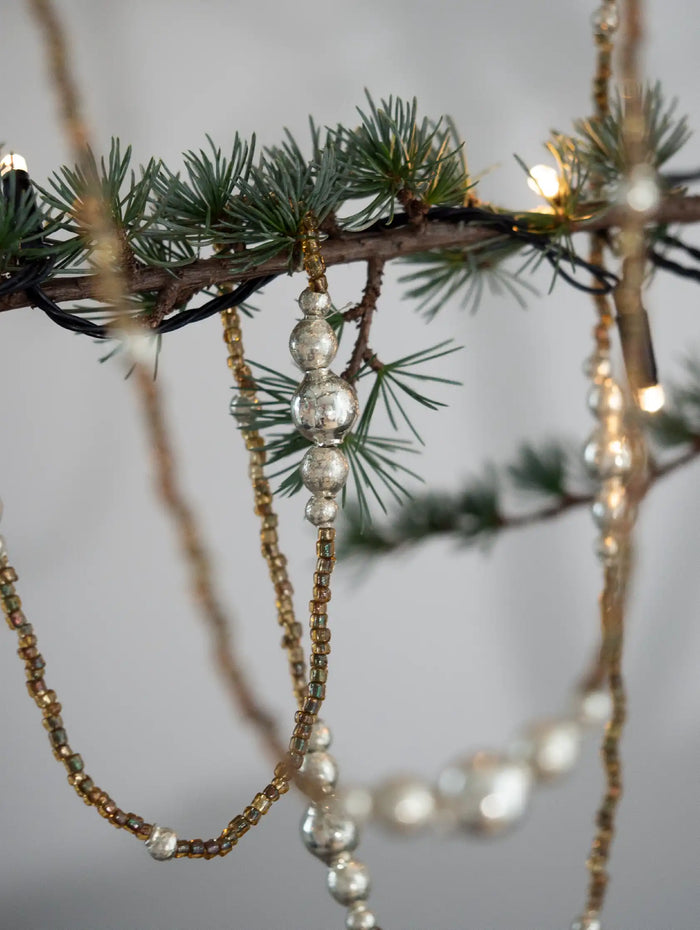 Decorative beaded strands hanging from a branch with small lights against a neutral background