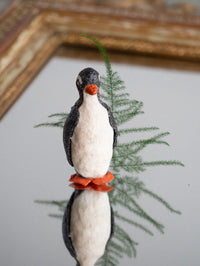 Felted penguin figurine with a red scarf on a branch against a neutral background