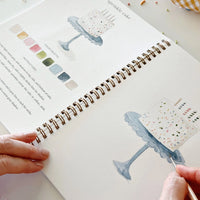 hand holding a brush and water coloring an illustration of a cake on a stand