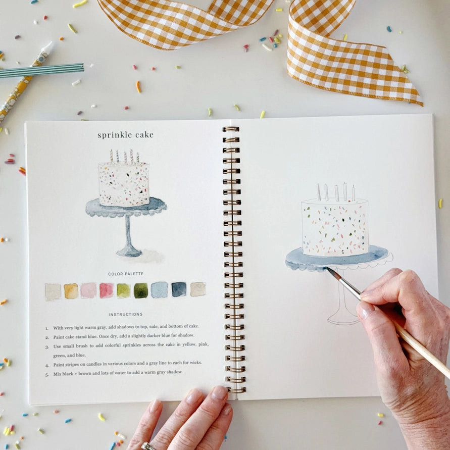 hand holding a brush and water coloring an illustration of a cake on a stand