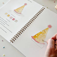 hand holding a brush and water coloring an illustration of a birthday celebration paper hat