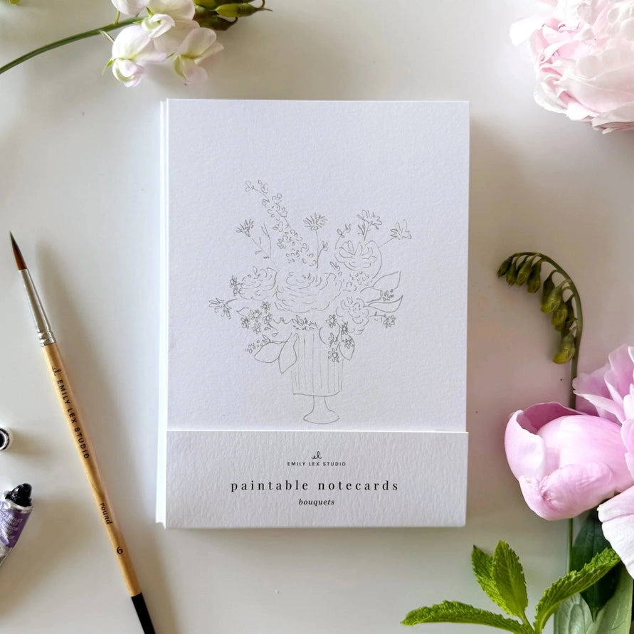notecard with an outline of a flower bouquet on a desk with a watercolor paint brush and flowers