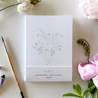 notecard with an outline of a flower bouquet on a desk with a watercolor paint brush and flowers