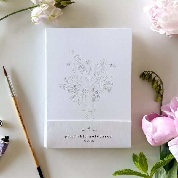 notecard with an outline of a flower bouquet on a desk with a watercolor paint brush and flowers