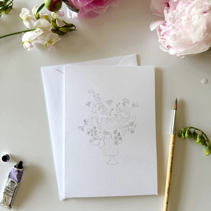 notecard with an outline of a flower bouquet on a desk with a watercolor paint brush and flowers