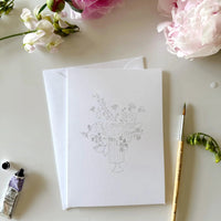 notecard with an outline of a flower bouquet on a desk with a watercolor paint brush and flowers