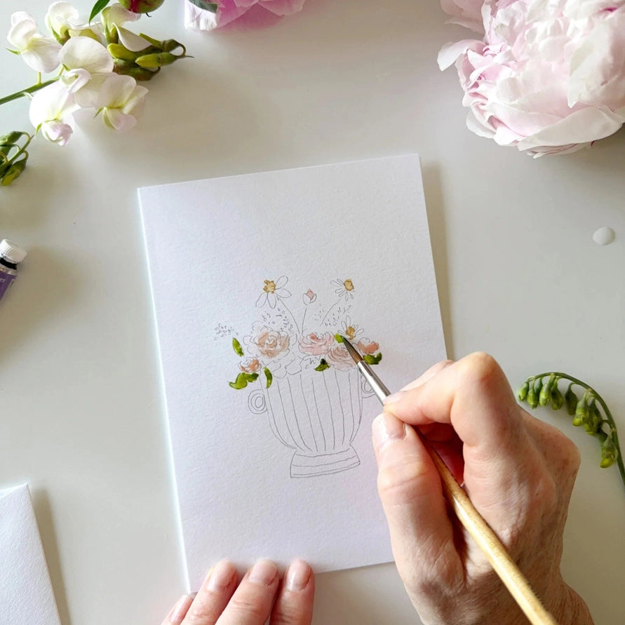 notecard with an outline of a flower bouquet on a desk with a watercolor paint brush and flowers