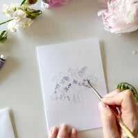 notecard with an outline of a flower bouquet on a desk with a watercolor paint brush and flowers