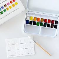 A watercolor paint set with a white metal case containing multiple color pans, shown open on a white surface. A paintbrush with a light wood handle rests in front, and part of the watercolor palette booklet is visible at the top left.