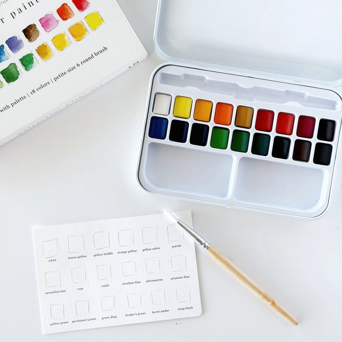A watercolor paint set with a white metal case containing multiple color pans, shown open on a white surface. A paintbrush with a light wood handle rests in front, and part of the watercolor palette booklet is visible at the top left.