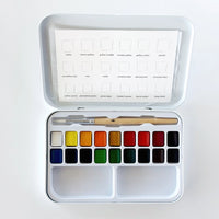 An open metal watercolor paint set with a white tray holding multiple color pans, a wooden-handled brush, and a sheet of color cards inside the lid, photographed from above on a white background.