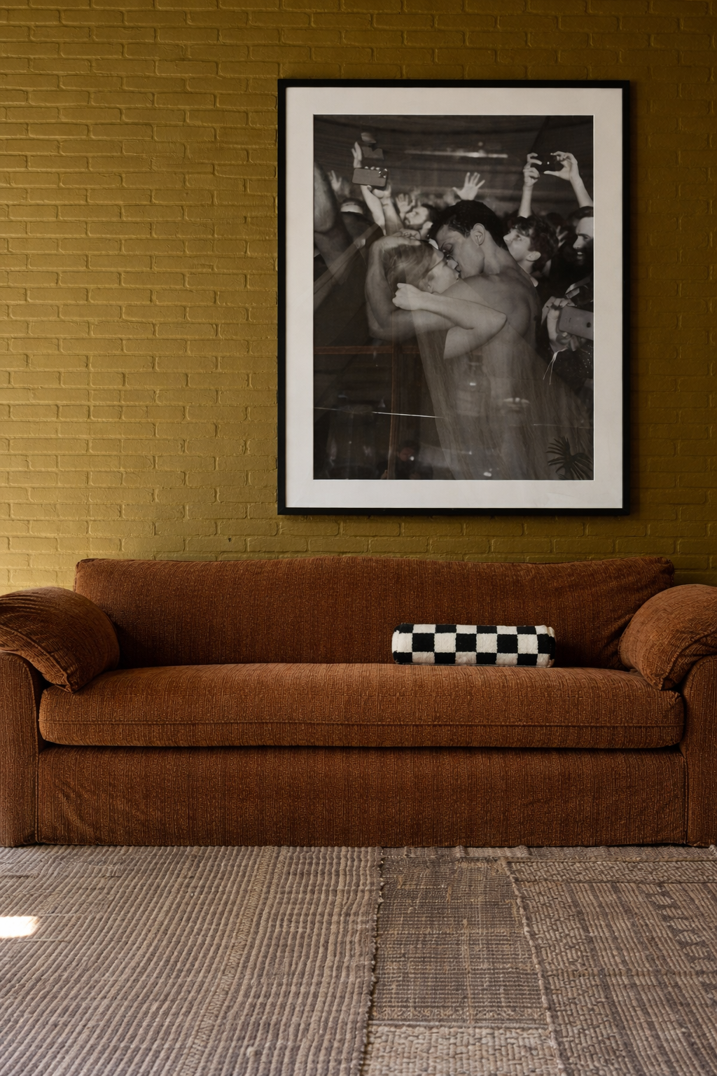 Brown sofa with a checkered pillow against a wall with a framed black and white photograph.