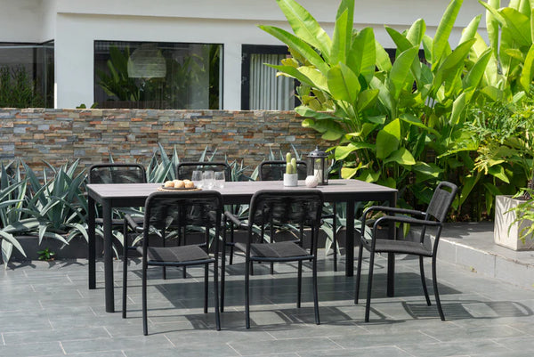Outdoor dining set with table and chairs on a patio area with plants and building in the background