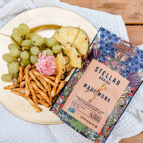 Stellar Braids Maui Monk package with a plate of food including grapes, pineapple, and fries on a wooden table.