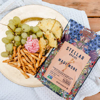 Stellar Braids Maui Monk package with a plate of food including grapes, pineapple, and fries on a wooden table.