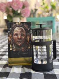 Coffee package with a face design and a French press on a checkered tablecloth.