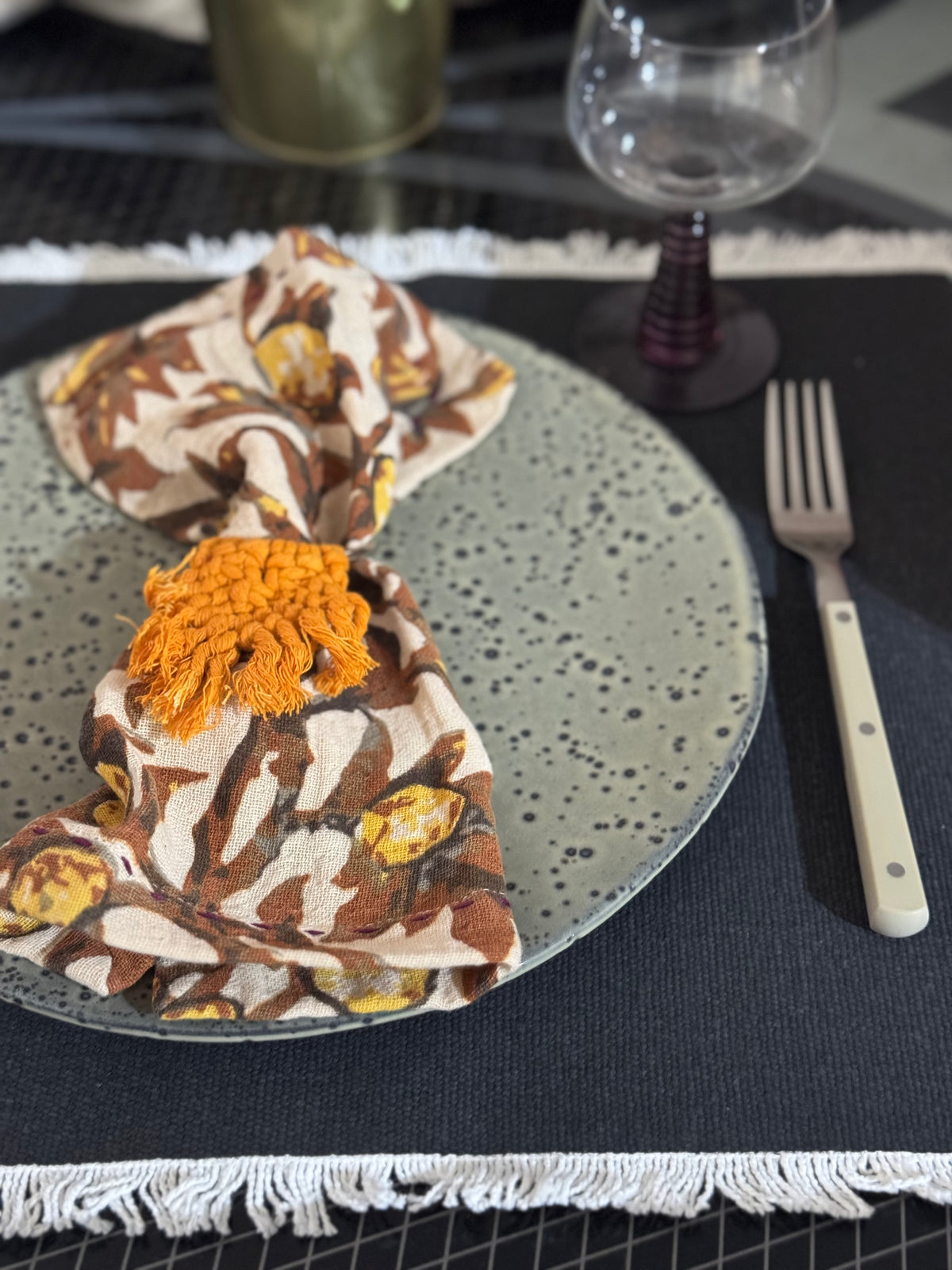Decorative napkin with orange pom-pom on a speckled plate with cutlery and glassware.