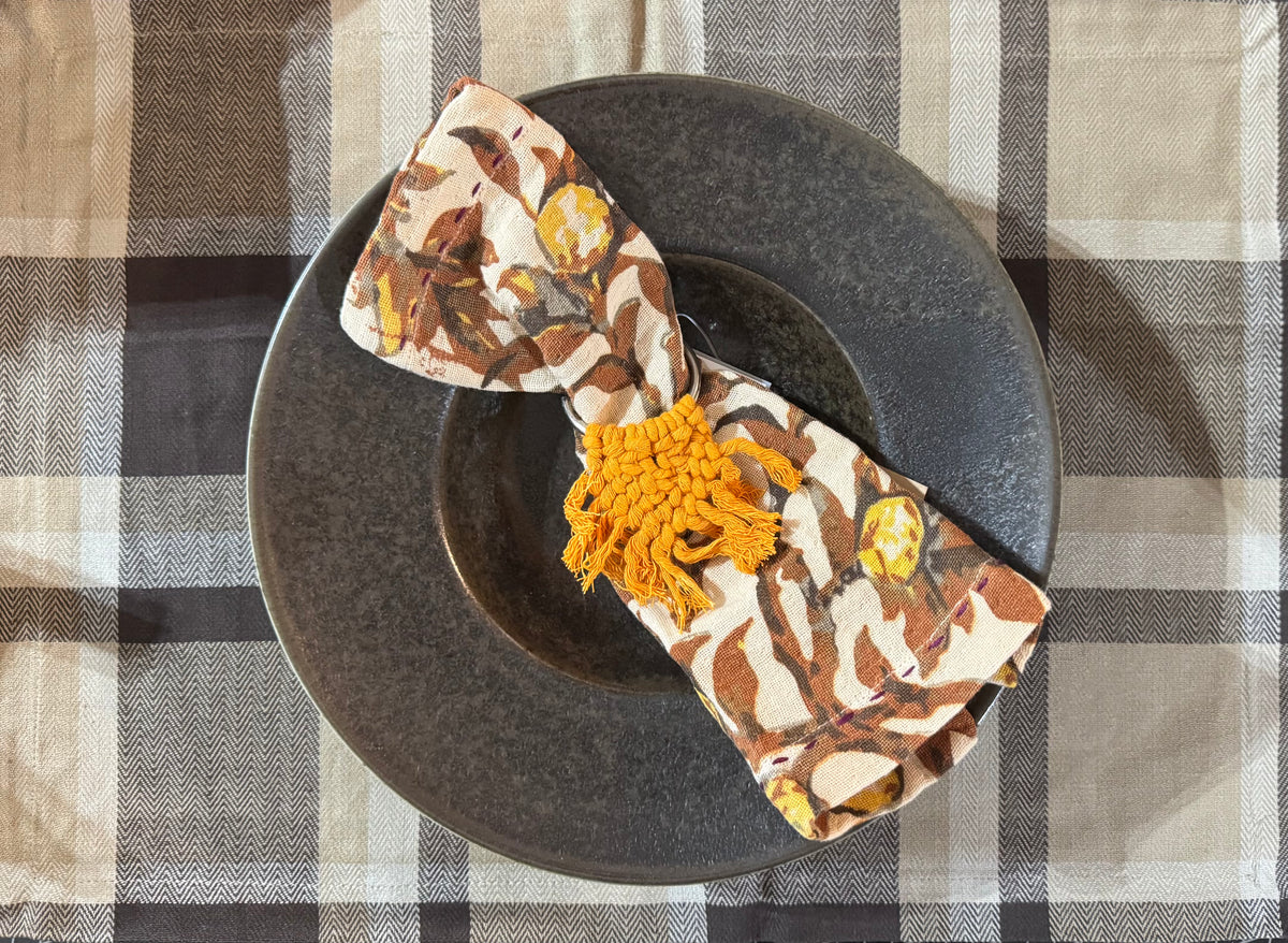Decorative napkin with floral pattern and orange tassels on a dark plate against a checkered tablecloth.