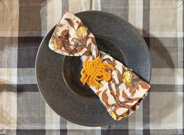 Decorative napkin with floral pattern and orange tassels on a dark plate against a checkered tablecloth.