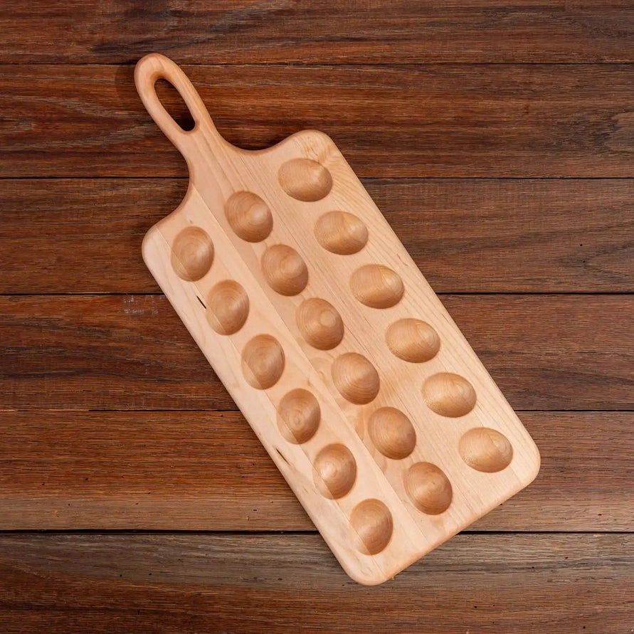 A rectangular wooden serving board with a loop handle and multiple oval depressions, photographed on a dark brown wood plank surface.