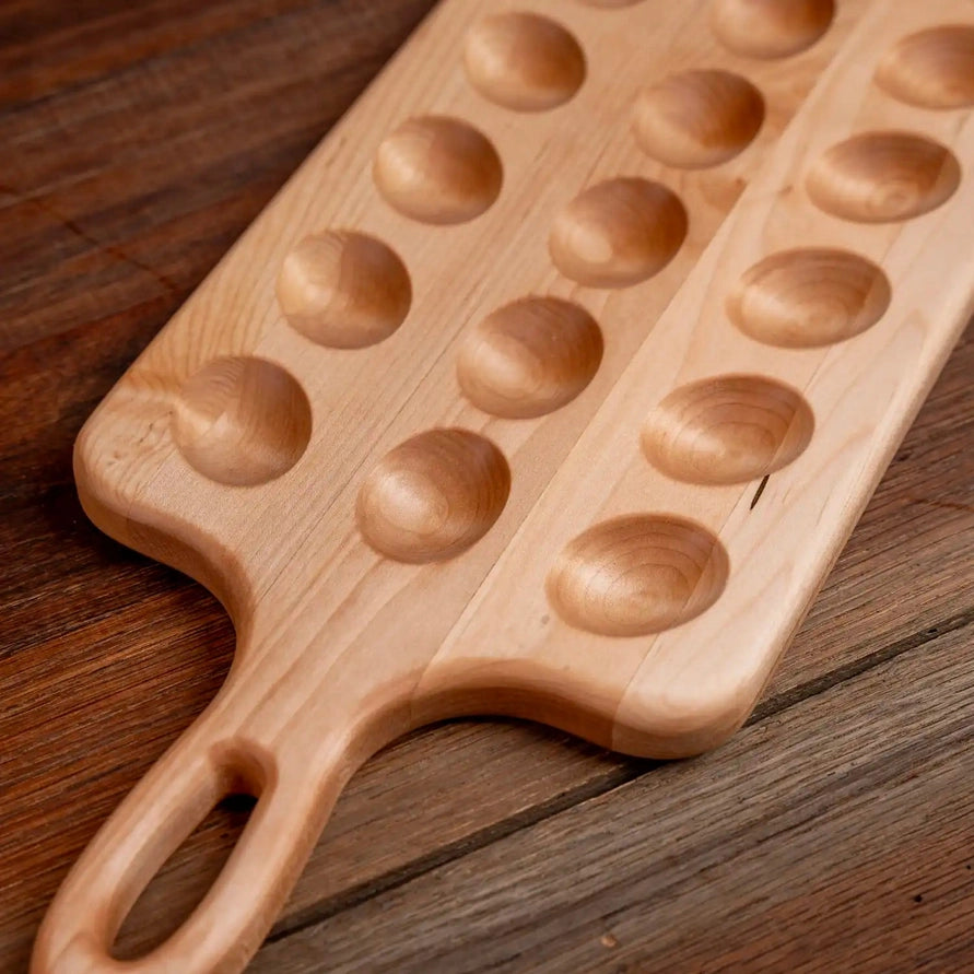A rectangular wooden serving board with a loop handle and three rows of oval recessed wells, photographed on a rustic dark wood surface.
