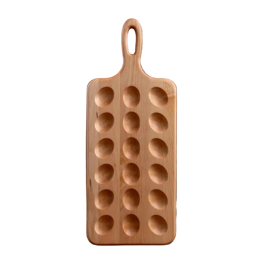 A rectangular wooden serving board with a loop handle and six oval cavities, standing upright on a wooden surface against a dark wood plank background.