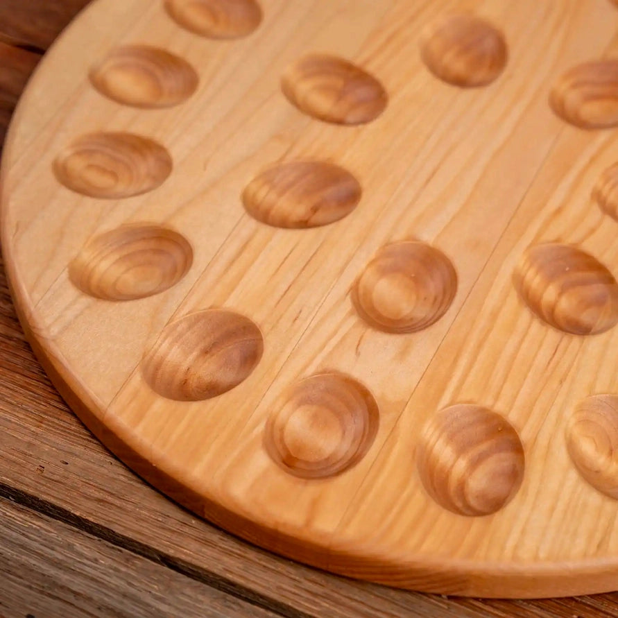 A close-up of a round light wood board with multiple oval recessed pits, photographed on a rustic wooden surface.