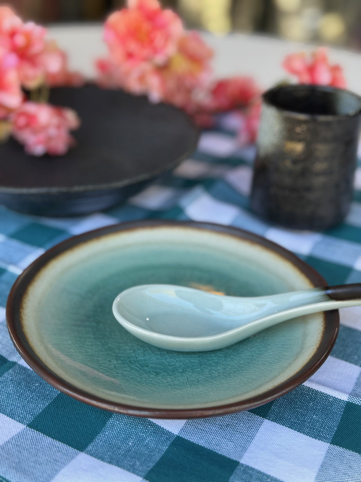 Teal ceramic plate with a spoon on a checkered tablecloth, with flowers and a vase in the background.