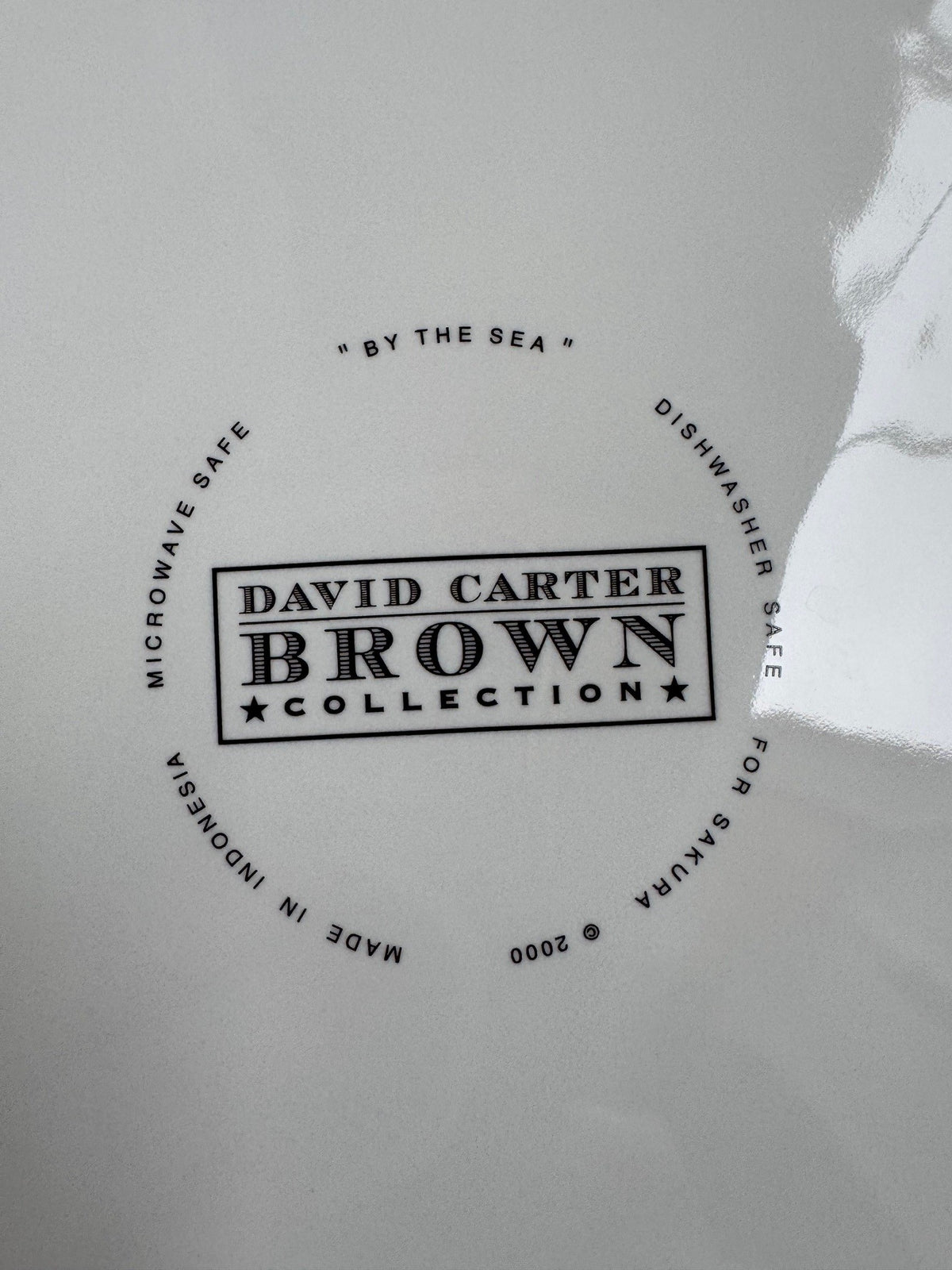 A close-up of a white glossy surface with a circular black printed logo reading 'DAVID CARTER BROWN COLLECTION' and surrounding text including 'MICROWAVE SAFE', 'DISHWASHER SAFE', and 'MADE IN INDONESIA'.