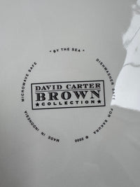 A close-up of a white glossy surface with a circular black printed logo reading 'DAVID CARTER BROWN COLLECTION' and surrounding text including 'MICROWAVE SAFE', 'DISHWASHER SAFE', and 'MADE IN INDONESIA'.