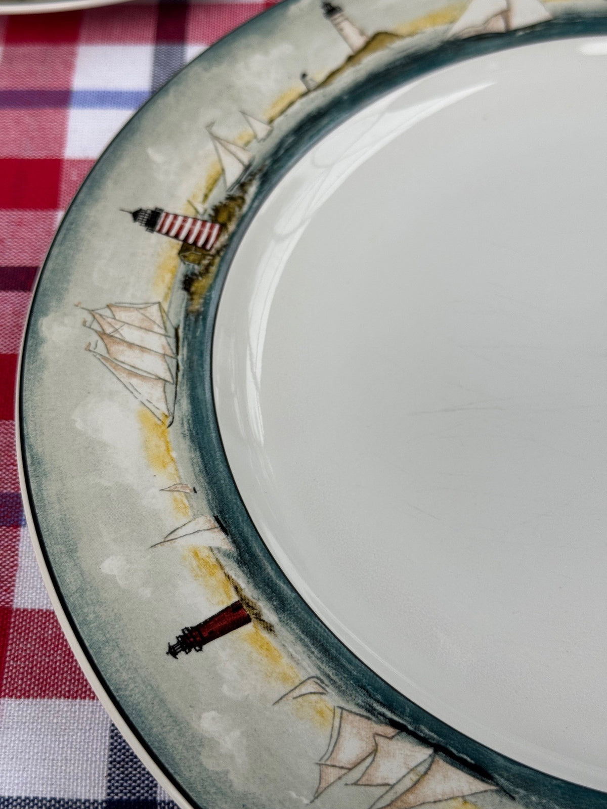 A close-up of a ceramic plate with a nautical design, featuring sailboats and lighthouses painted along the rim, set on a red and white checkered tablecloth.