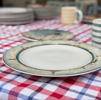 A ceramic dinner plate with a white center and a green, blue, and gold patterned rim, set on a red, white, and blue checkered tablecloth. Other plates and striped mugs are blurred in the background.