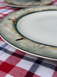 A close-up of a ceramic plate with a white center and a painted rim featuring sailboats and small boats in muted green, gray, yellow, and red tones, resting on a red, white, and navy checkered tablecloth.
