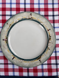A round ceramic plate with a lighthouse and sailboat border design, placed on a red, white, and blue plaid tablecloth.