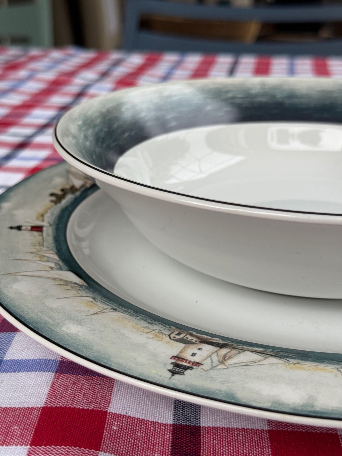A white ceramic bowl with a dark blue-gray rim sits on a matching plate featuring a lighthouse illustration, set on a red and white checkered tablecloth.