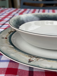 A white ceramic bowl with a dark blue-gray rim sits on a matching plate featuring a lighthouse illustration, set on a red and white checkered tablecloth.