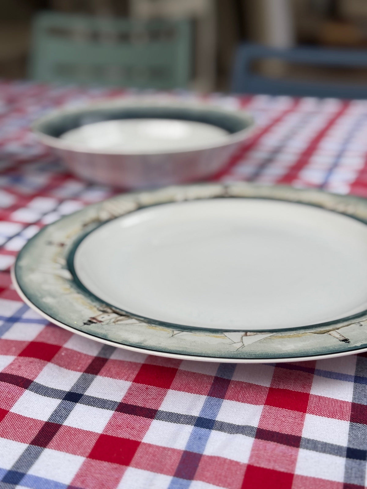 A ceramic plate with a white center and a green-beige patterned rim sits on a red, white, and blue checkered tablecloth, with a matching bowl blurred in the background.