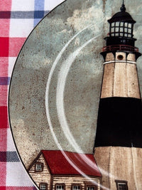 A close-up of a decorative plate featuring a lighthouse and a small house, placed over a red, white, and blue plaid fabric background.