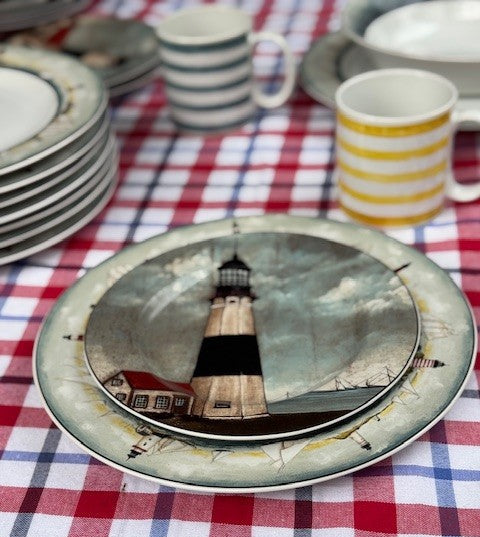 A stack of ceramic plates featuring a lighthouse illustration, set on a red, white, and blue checkered tablecloth alongside striped mugs.