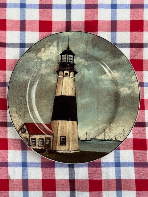 A decorative ceramic plate featuring a painted lighthouse scene, placed on a red, white, and blue checkered tablecloth.