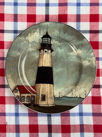 A decorative ceramic plate featuring a painted lighthouse scene, placed on a red, white, and blue checkered tablecloth.