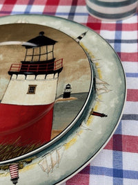 A close-up of a decorative ceramic plate featuring a painted lighthouse scene, set on a red, white, and blue checkered tablecloth.