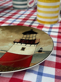 A decorative ceramic plate featuring a painted lighthouse design, placed on a red, white, and blue checkered tablecloth with striped mugs in the background.
