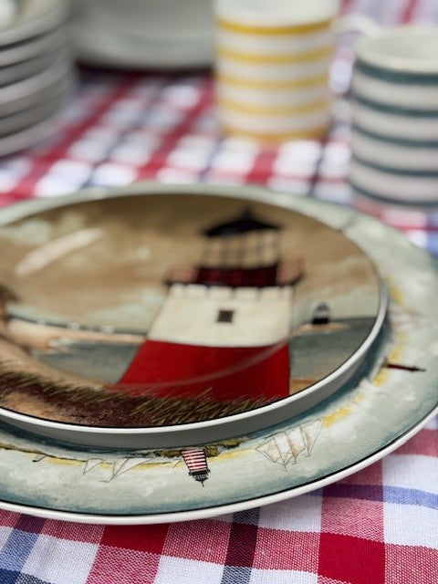 A close-up of a decorative ceramic plate featuring a red and white lighthouse illustration, stacked on a matching plate over a red, white, and blue checkered tablecloth.
