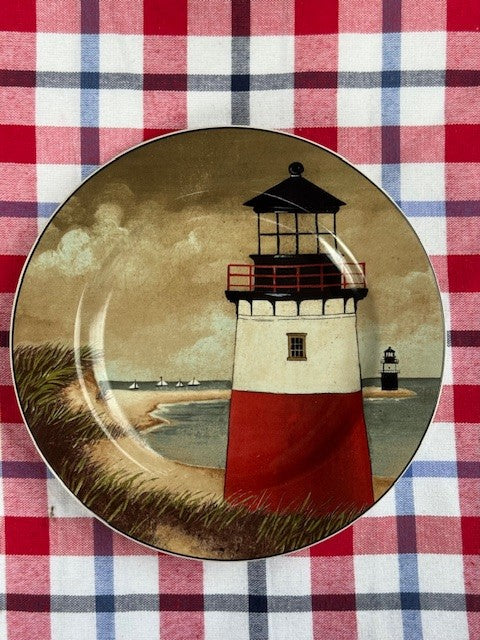 A decorative ceramic plate featuring an illustration of a red and white lighthouse on a beach under a cloudy sky, set against a red, white, and blue plaid fabric background.