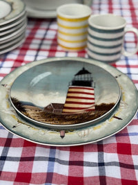 A decorative ceramic plate featuring a lighthouse scene, stacked on top of a matching plate over a red, white, and blue checkered tablecloth, with striped mugs in the background.